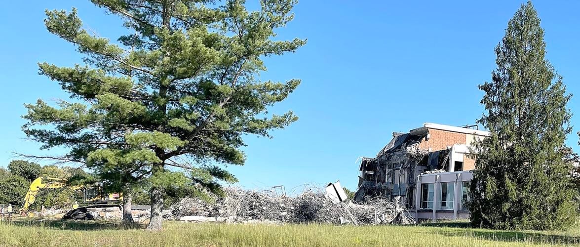 green valley demolished Under a clear blue sky, an excavator and mounds of twisted rebar and concrete surround a half-demolished brick facility at the former Greene Valley Developmental Center, framed by two tall evergreens.