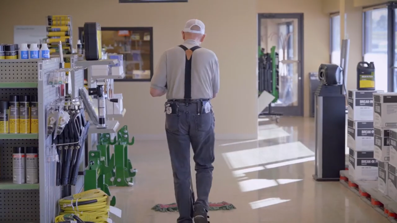 A rear view of Danny walking down a well-lit store aisle. He's in the same outfit, with tools and accessories clipped to his belt, surrounded by hardware and agricultural supplies.