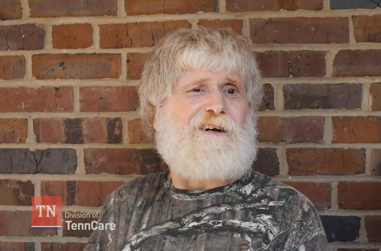 Donald, an older white man with light skin, a full white beard, and thick, light gray hair, smiles while standing in front of a brick wall. He is wearing a camouflage-patterned shirt. The Tennessee Division of TennCare logo appears in the bottom left corner.