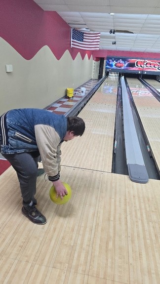 A man with fair skin and short brown hair, wearing a blue and gray varsity-style jacket, bends down to roll a bright yellow bowling ball at Kingpin Lanes. The bowling alley features red and beige walls, an American flag hanging above the lanes, and a ramp positioned for accessibility.