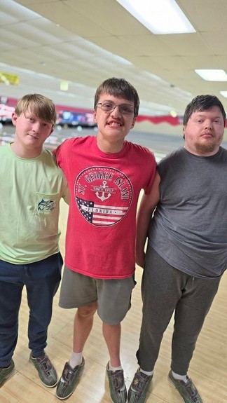 Three young men stand close together on a bowling lane, smiling and relaxed. From left to right: one wears a light green shirt and navy pants, the next wears a red “St. George Island” sleeveless T-shirt and glasses, and the third wears a gray T-shirt and matching pants. All three are wearing bowling shoes, enjoying their Guy’s Night Out at Kingpin Lanes.