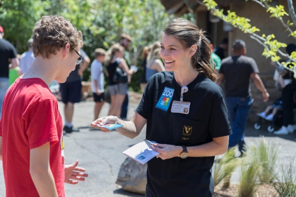 A smiling, light-skinned woman with a brown ponytail is wearing a Vanderbilt TRIAD shirt with a Nashville Zoo sticker. She is handing a similar sticker to a young man with light skin, wearing a red shirt. A line of people is visible in the background. 