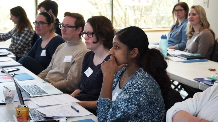 Two rows of adults sit at tables in a windowed classroom setting, laptops and papers in front of them, their attention focused toward the front of the room.