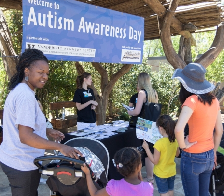 Two women – one dark skinned, the other light skinned with – with young girls and strollers stand in front of an exhibit table under a wooden canopy with a sign that reads, “Welcome to Autism Awareness Day.” The Vanderbilt Kennedy Center and Predators Foundation logos are visible on the sign. A young woman stands behind the table engaging with a blonde woman holding printed materials. 