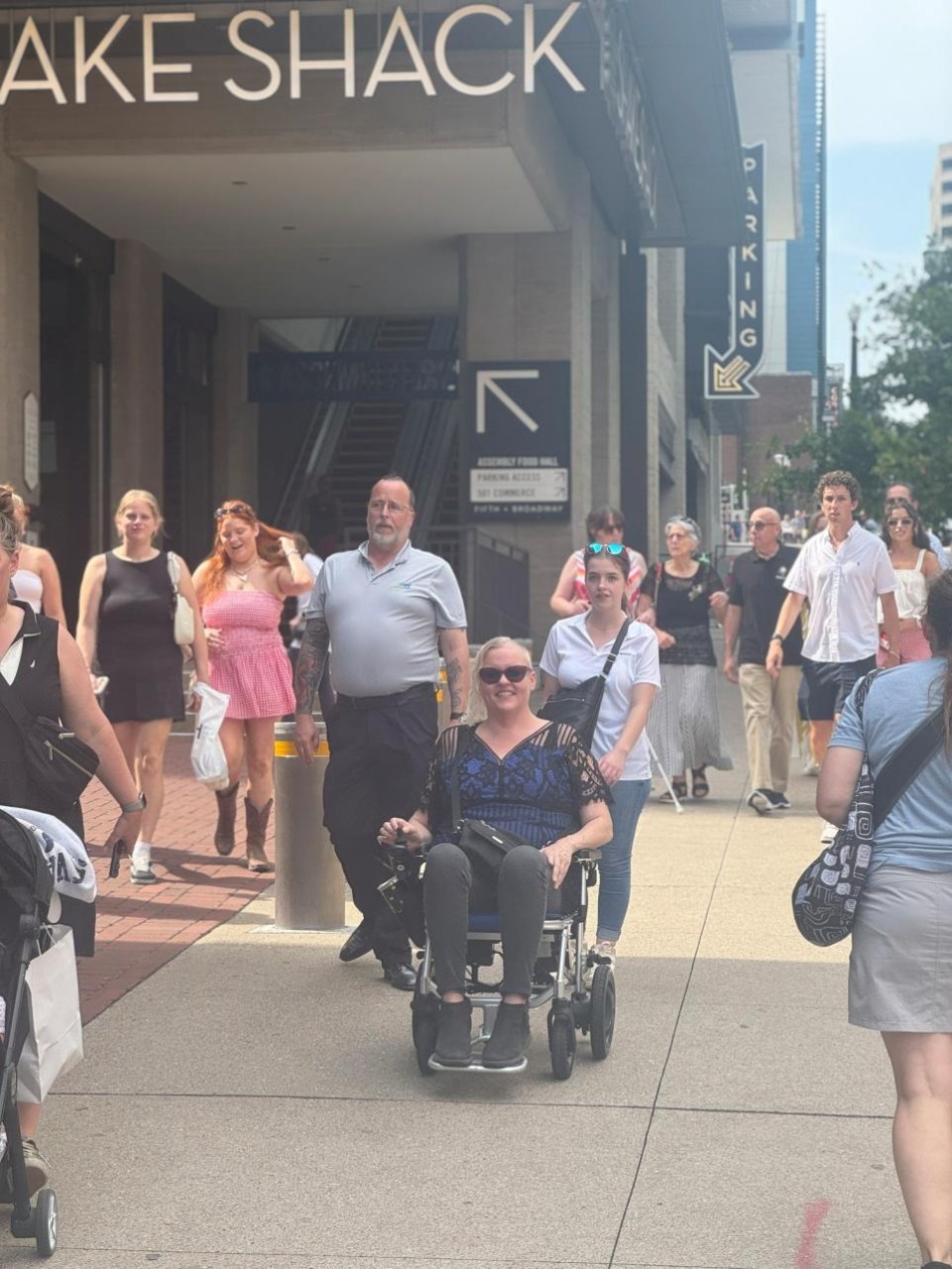 Cynthia George, a fair-skinned woman with platinum-blonde hair, is smiling while using a power wheelchair on a busy sidewalk outside the 5th and Broadway multiplex in Nashville. She wears sunglasses, a dark top with sheer lace sleeves, and dark pants. A crowd of people walks around her near the Shake Shack entrance.
