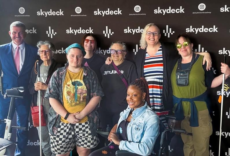 A group of people pose in front of a black "skydeck" step-and-repeat backdrop. In the front row, Keona Gwinn, a Black woman with braided hair, sits in a wheelchair wearing a black top and a light denim jacket. To her left, Dirk Strider, a white person with short hair and a teal bandana wears a tropical shirt and checkered shorts. In the back row, Cynthia George, a fair-skinned woman with platinum-blonde hair and glasses, stands smiling in a black cardigan over a red, white, and blue striped dress.