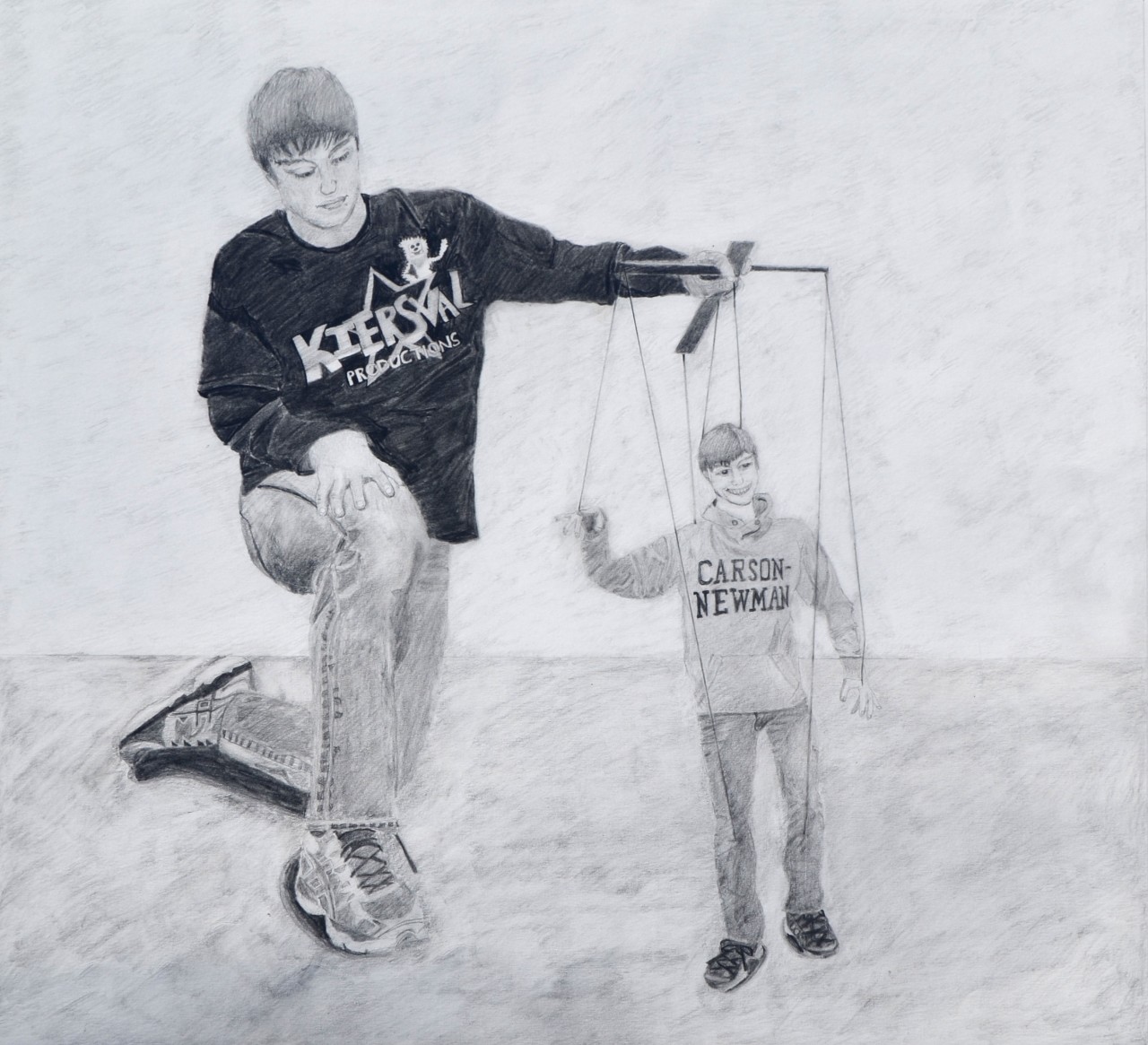  a detailed pencil drawing by Kieran Braun called, “Manipulation”. It features a young man in jeans and a dark t-shirt that has the word “Kiersval Productions” written across it. He is kneeling and holding a marionette on a frame with strings. The marionette is a boy wearing a Carson-Newman sweatshirt.