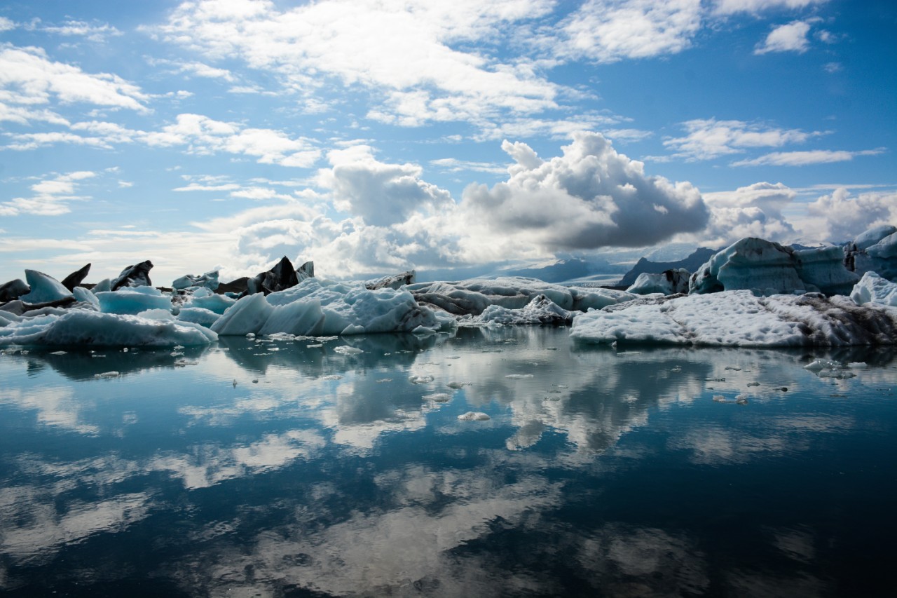 photo called Glacier Lagoon, and shows white ice caps on a shiny body of water, with an expansive blue sky with clouds above it. You can see the reflections of the sky, the clouds and the ice caps in the surface of the water. 