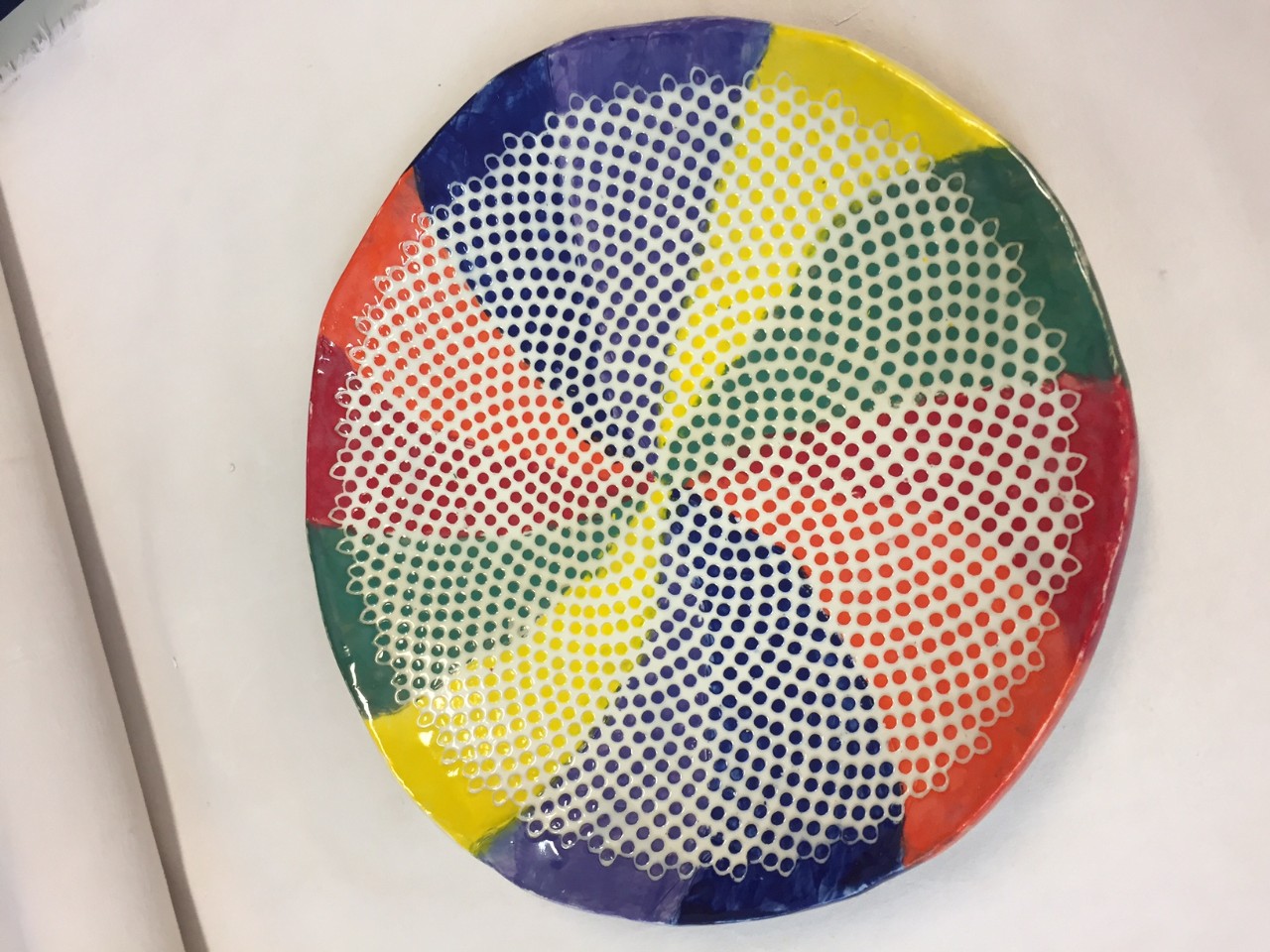 a photo of a beautiful ceramic plate that is called, “Polka Dot Pinwheel. The pottery is by Louise McKown. The surface of the plate is divided into 12 slivers. Two are yellow, two are orange, two are red, two are green, two are dark blue and two are light blue. In each of the slivers, as you move toward the center of the plate, the solid colors described above are replaced with many dots in those same colors. 