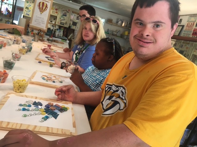 our people sitting along a long table working on their mosaic design components. The closest young man has Down syndrome and he is smiling and looking directly into the camera