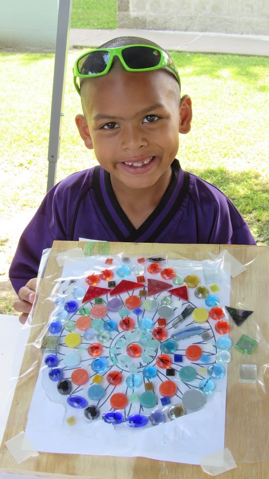 a young boy holding up his completed part of the mosaic design. He is wearing a purple sports jersey and there is a pair of green sunglasses pushed up onto his forehead.