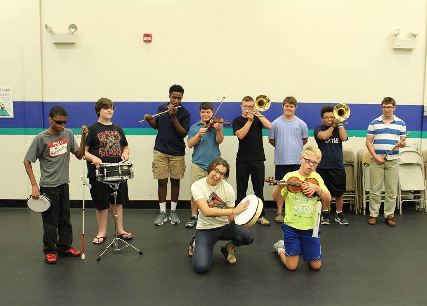 a group of boys, each holding a different instrument, with a female and male instructor kneeling on the ground in front of them