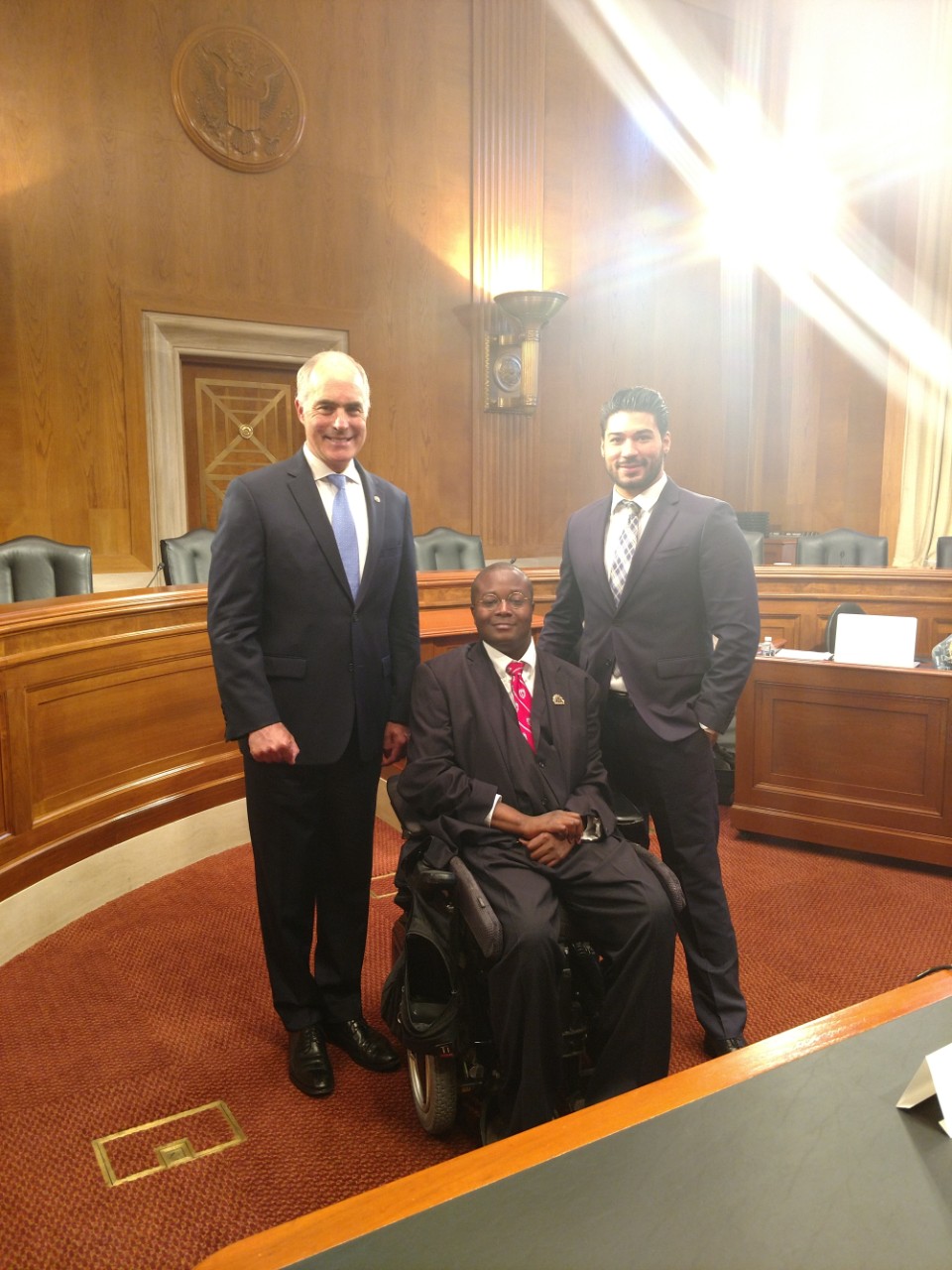 There are three men posing in a federal government meeting room in Washington, DC. They are Senator Bob Casey, Christopher J. Rodriguez, Director of Public Policy, and Edward Mitchell, the subject of the article. All three men are wearing suits and ties, and Edward Mitchell is sitting in a power wheelchair.