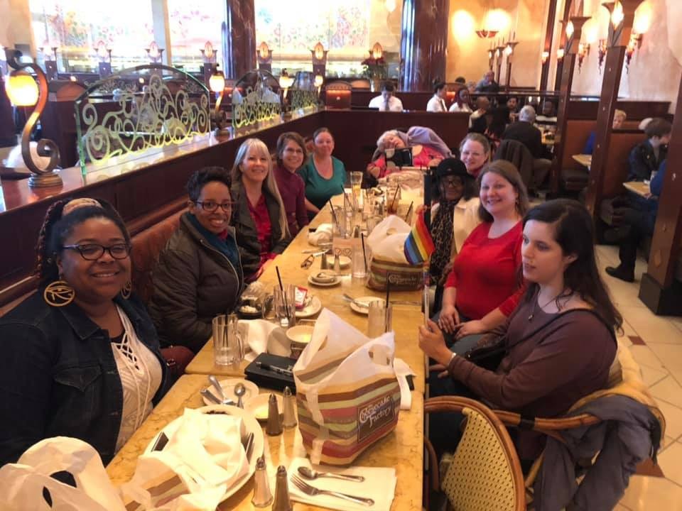 From left to right, circling around a long rectangular table in a Cheesecake restaurant: Ebony Cole, Clarisse Durnell, Laura Cox, Britt Cox, Rachel Batey, Lorri Mabry, Nicole Worthy, Albrice Alred, April Meredith and Becky Scott.” The table is covered with Cheesecake Factory to go bags, drink glasses with straws, and plates.