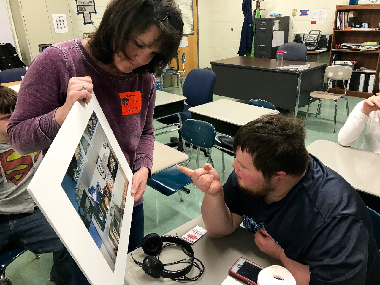 Author of the article Jen Vogus holds up a framed photo in a classroom and the young adult student seated in front of her is leaning forward and pointing at it