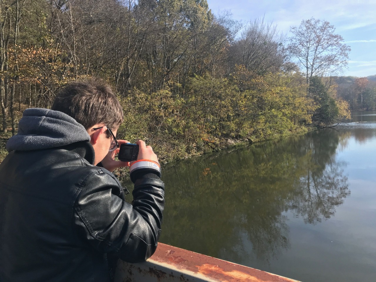 a young man stands on a bridge over a lake, with trees on the bank - he is taking a photo and wearing sunglasses and a windbreaker on a sunny day