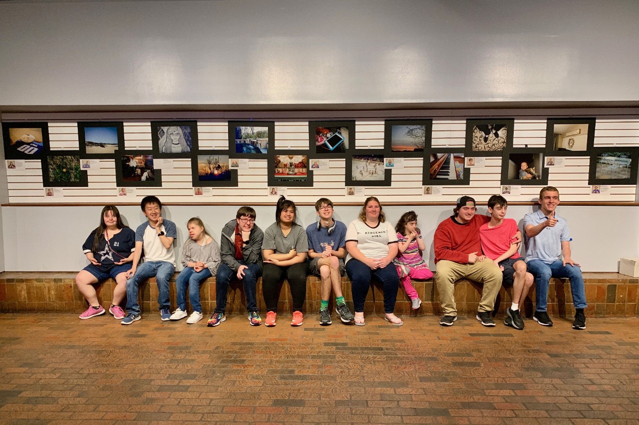 a group of about a dozen young adults with disabilities are seated in an art gallery on a bench; above them hang framed photographs taken by the students