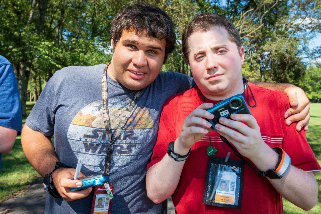 two young men with disabilities stand together with their arms around each other and holding small cameras during an AbleVoices activity outside