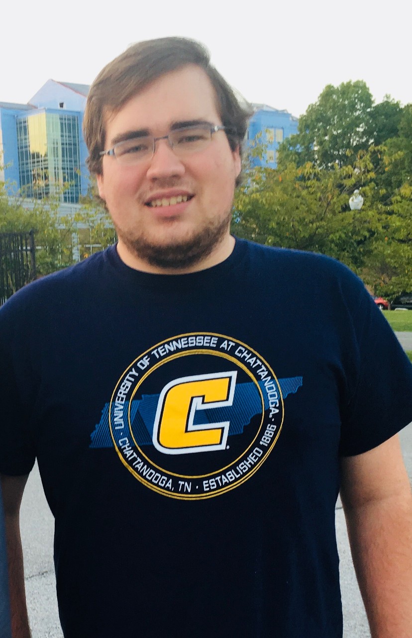 a young man with brown hair and a little bit of a beard in a university of tn chattanooga black t-shirt standing outside