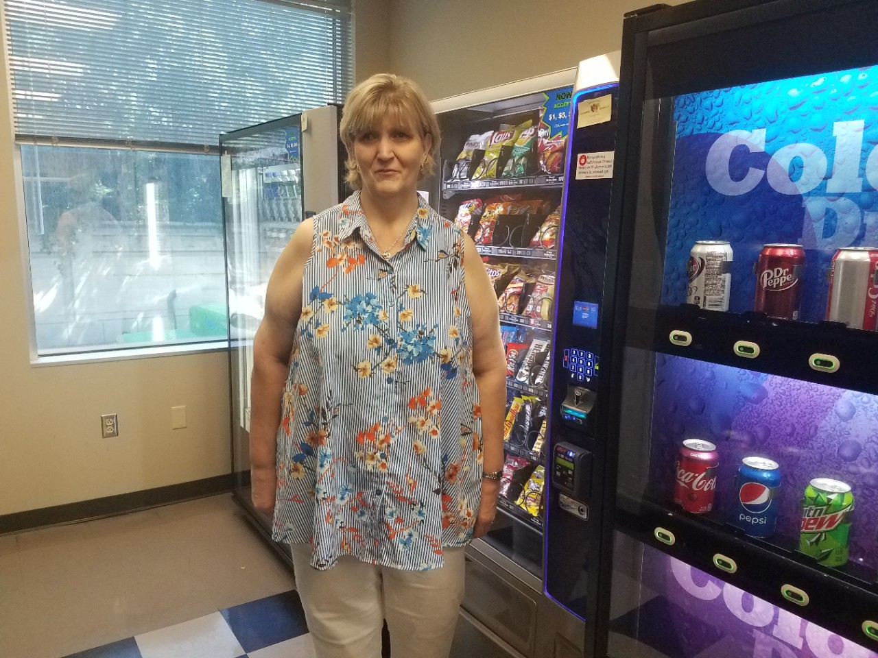 a blonde woman in a flowery sleeveless shirt stands in the break room and cafeteria of the state office building where she works, with vending machines behind her