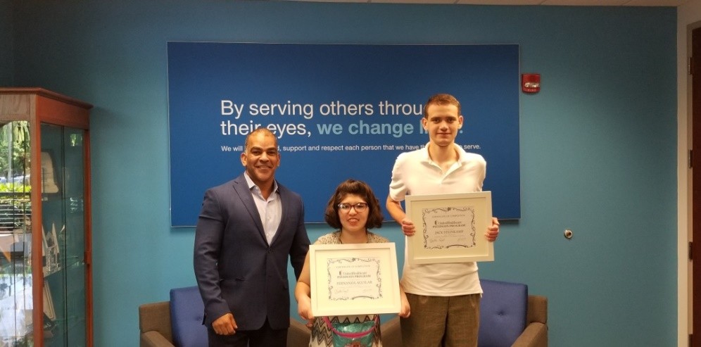 two young adults with disabilities hold up certificates and stand next to a gentleman in a suit in the lobby of an office