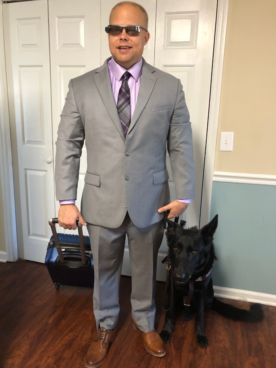 picture of article author James Brown standing in front of a closet door with his black assistant dog, Vantis. James is very professionally dressed in a gray business suit, and he is pulling a blue suitcase on wheels. His other hand holds his dog’s harness.