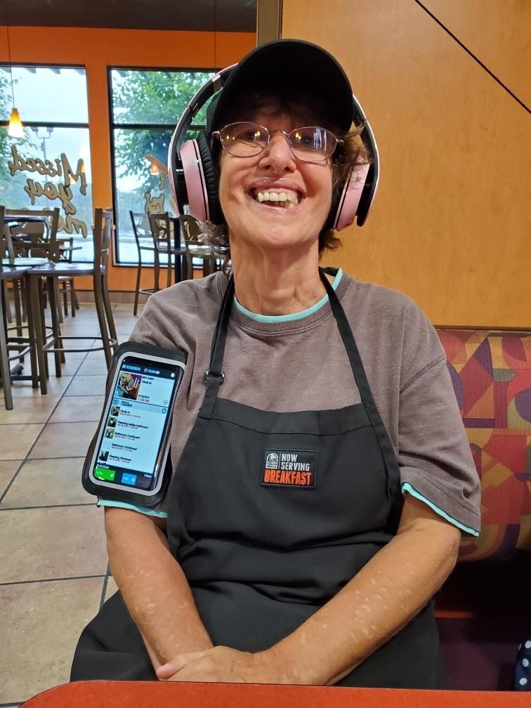 a photo of Anita - a close-up of a woman working in a Taco Bell restaurant. She is wearing an apron over a gray sweatshirt. The apron has a badge with a Taco Bell company logo and the words, “now serving breakfast.” The employee also has a smart phone strapped to her right arm, and she is wearing large, pink headphones over her ears. She wears glasses, and has a huge smile. In the background you can see several customer tables with empty chairs, and the large, front windows