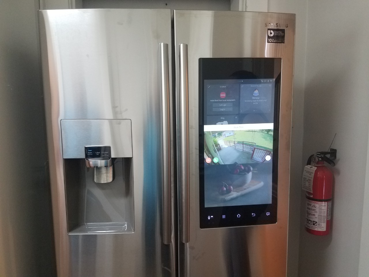 a photo of a stainless steel refrigerator with a large digital panel in one of the doors that shows the view outside the front door from a camera, and has other features where a person can make shopping lists, use a calendar and many many other tech functions