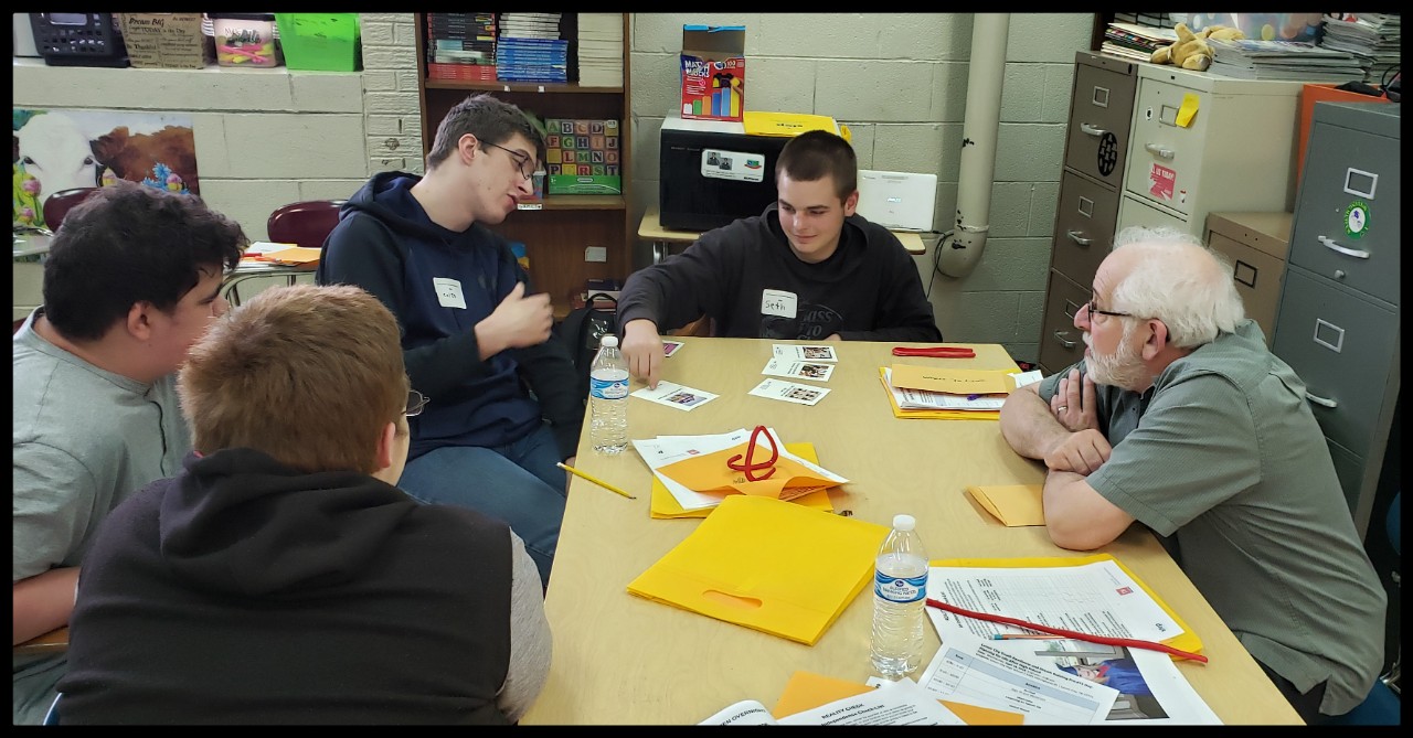 Ned at a recent Youth Readiness Day training for high school students with disabilities; he sits with 3 young men with disabilities at a table in a school classroom as they do an activity and discuss things together