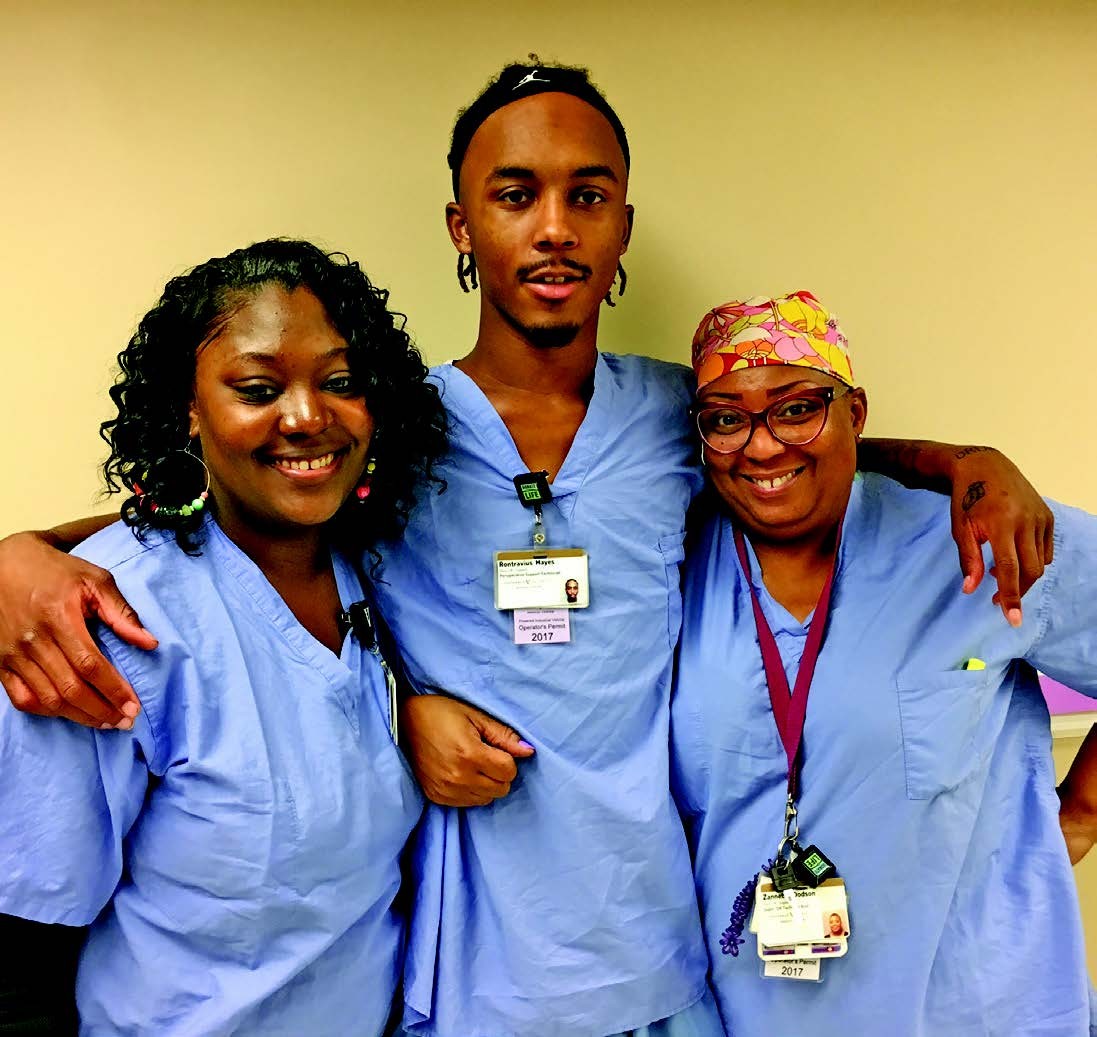 three African American people, co-workers, with arms around each other. They are all wearing blue hospital scrubs. They are, left to right, Raykeyda, Ronand Ms. Z. 