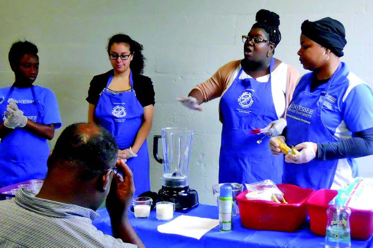 four TSU staff members in blue aprons are involved in a cooking class. They are from left to right: Shanail Brown (TSU SNAP-Ed PA, Davidson), Marina Fahmy (Intern, Davidson), Ebone’ Colclough (SNAP-Ed Agent, Davidson) and Latasha Holloway (EFNEP PA, Davidson).
