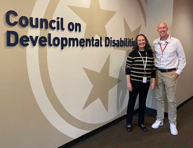 Two people are standing in front of a wall with the text "Council on Developmental Disabilities" and a large design featuring three stars. On the left, Jolene Sharp, a woman with a light skin tone, has long, dark, wavy hair and is wearing a black sweater with beige stripes, black pants, and black shoes. She has an ID badge on a white lanyard. On the right, Blake Shearer, a man with a light skin tone and a bald head, is wearing a white and red checkered shirt, beige pants, and white sneakers. He has an ID badge on a red lanyard. Both are smiling and standing side by side.