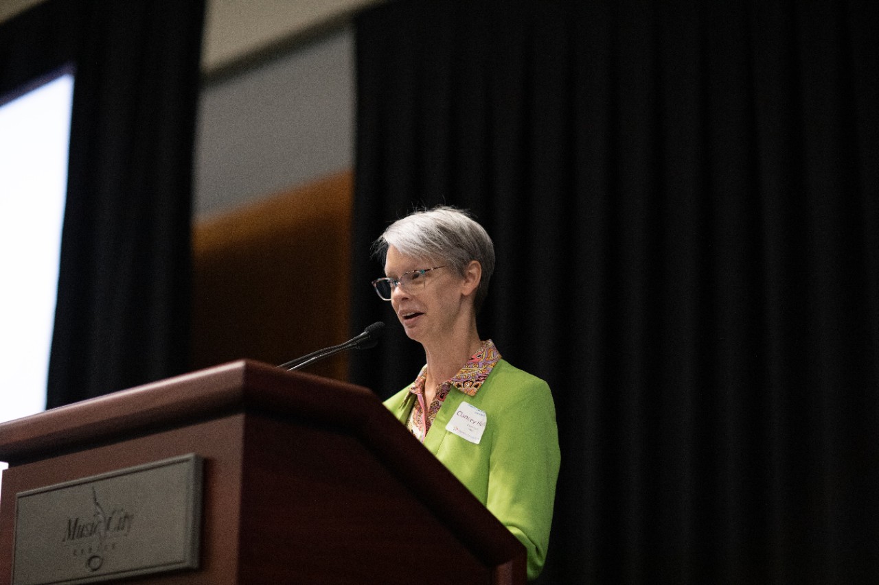 Clancy Hopper, a light-skinned woman with short gray hair and glasses, speaking at a podium. She is wearing a bright green blazer with a patterned shirt underneath and is addressing an audience, standing in front of a dark curtain.