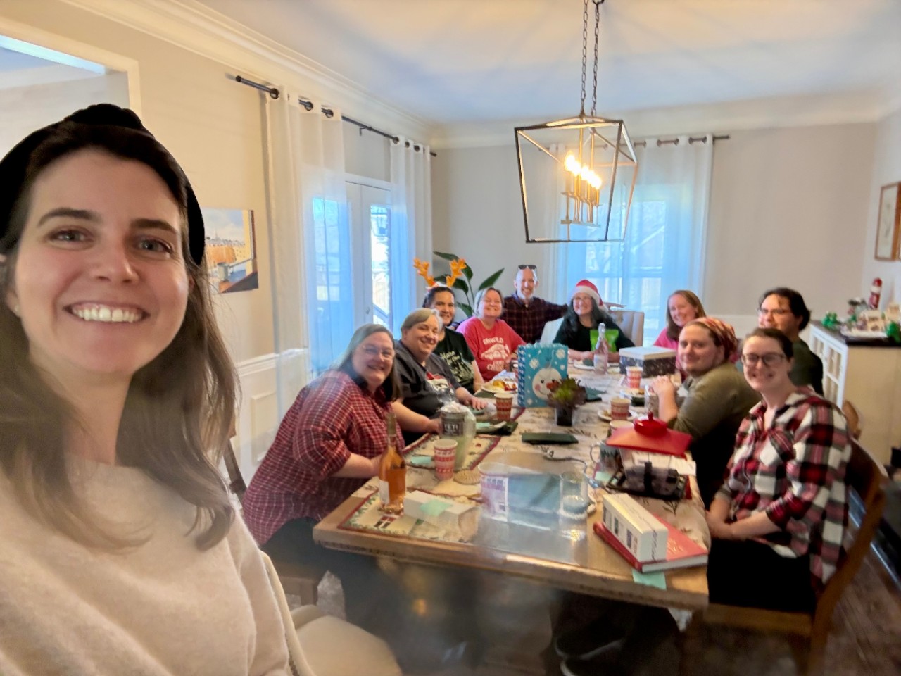 Group photo of eleven people smiling around a dining table at a holiday gathering. One person in the foreground takes a selfie than includes the others who sit with drinks, gifts, and notebooks. The room is bright with large windows, white curtains, and a modern hanging light, creating a warm, festive atmosphere.