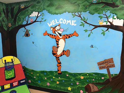 Photo of wall painting in a child visitaiton area.  Painting has trees, tiger, owls, bubble bees, flowers, and a sign that says Hundred Acre Woods.