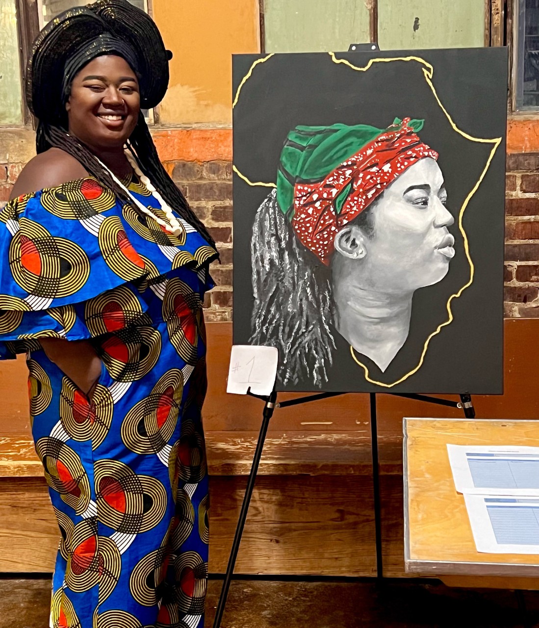 a woman in a colorful outfit stands next to a painting and portrait she created, smiling