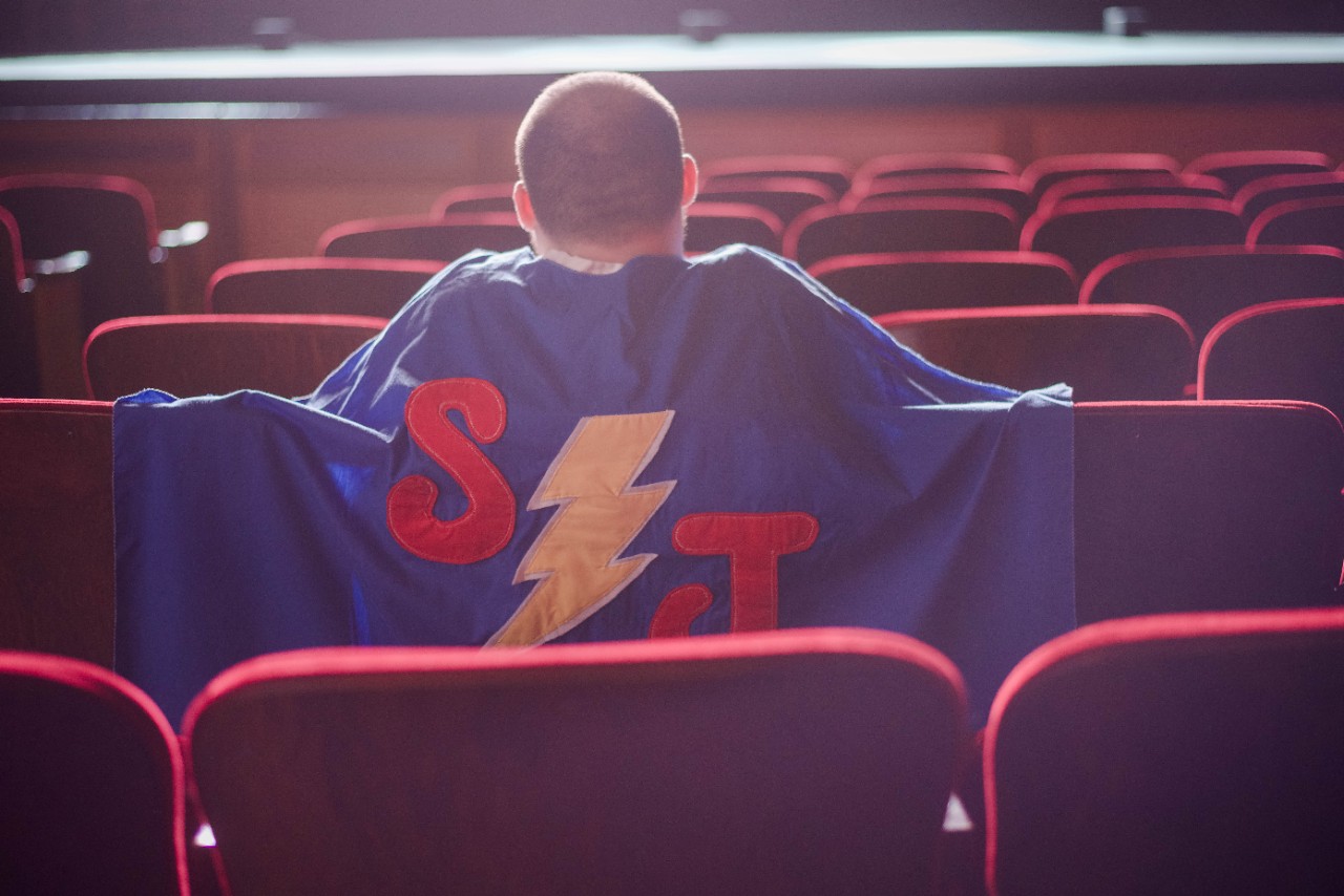 JacobintheSeats Jacob sits in rows of red theatre chairs with his back to the camera. His blue JS cape drapes over the back