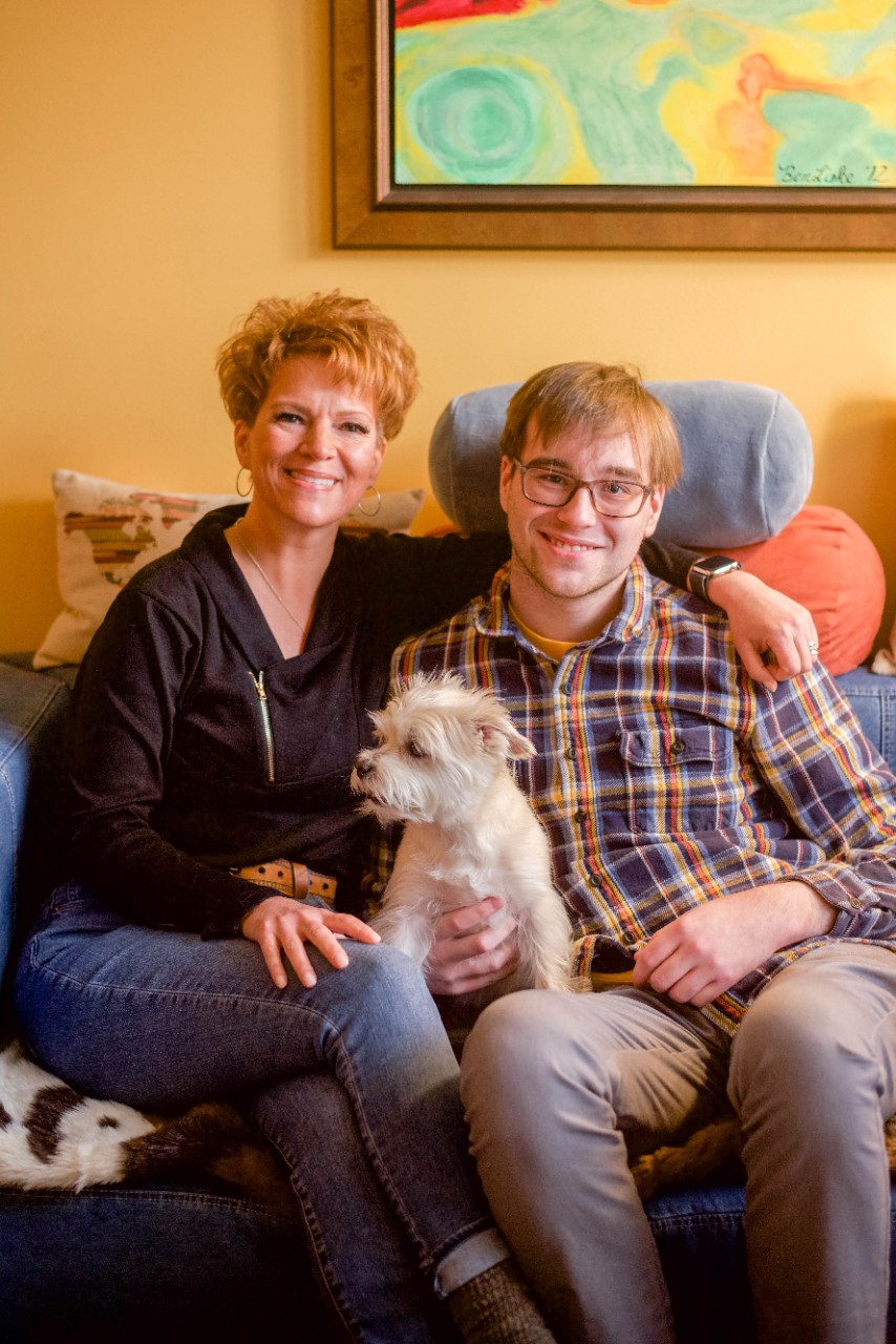 Juli, a 51 year old woman with light skin and short red hair, sits with Ben, her 21yo son, a man with light skin and short dark hair with glasses. Their small white dog sits in their lap on the living room couch