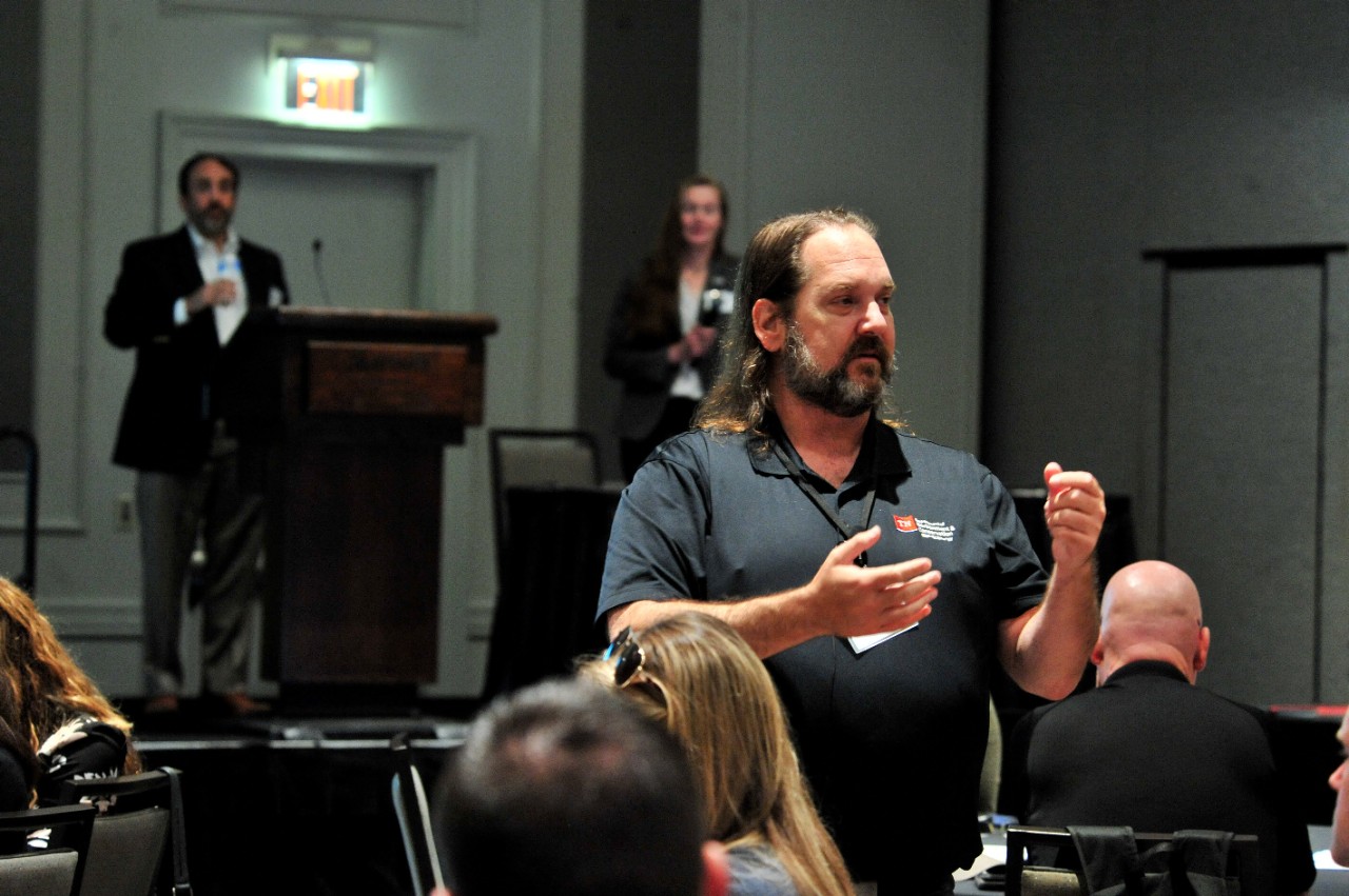 Ben Bolton, Assistant Director of Energy Security, Emergency Preparedness, and Resilience, standing while facilitating the Operation Catfish tabletop exercise in a ballroom of people sitting a round tables. In the background, a male speaker in a sport coat stands at a podium.