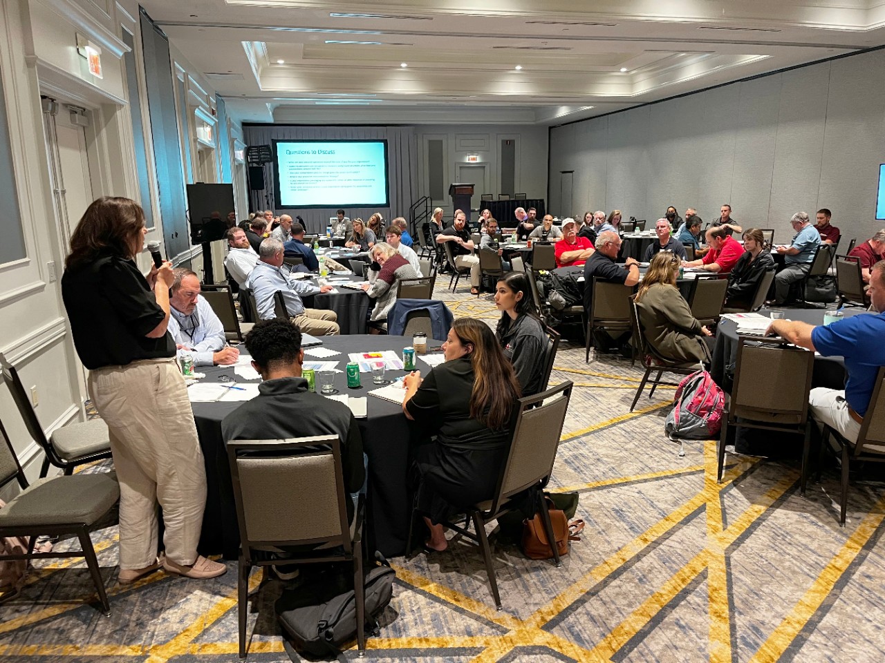 In the far left corner, a female exercise participant has her back to us as she reports out her table’s discussion to exercise participants sitting at round tables in a conference ballroom.