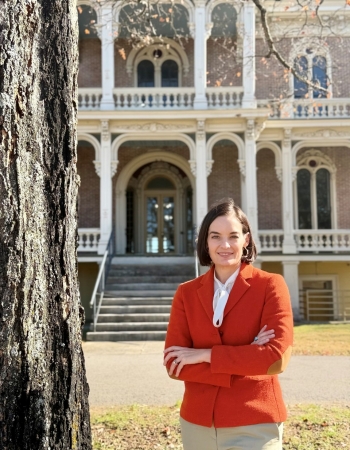 Image of woman in an orange jacket standing next to a tree with a historic home in the background