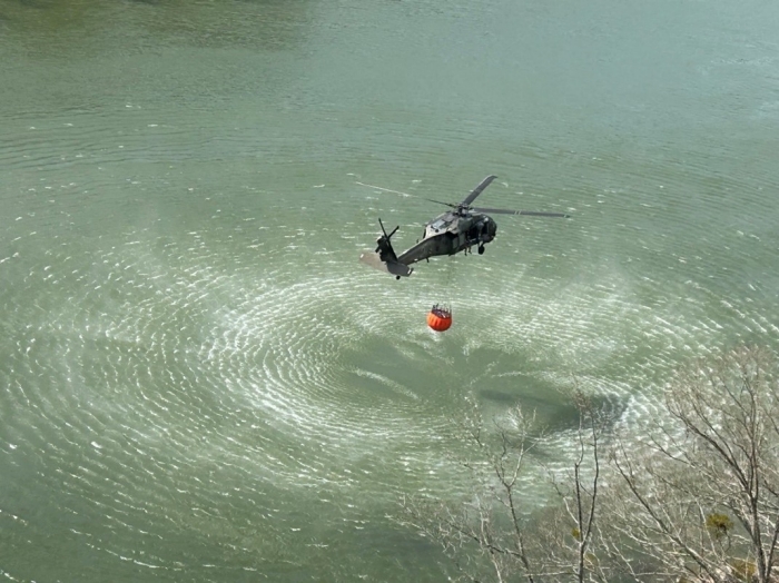 A Tennessee National Guard Blackhawk Helicopter hovers over a large body of water using a large bucket hanging from the helicopter to get water. A Tennessee National Guard Blackhawk Helicopter hovers over a large body of water using a large bucket hanging from the helicopter to get water.
