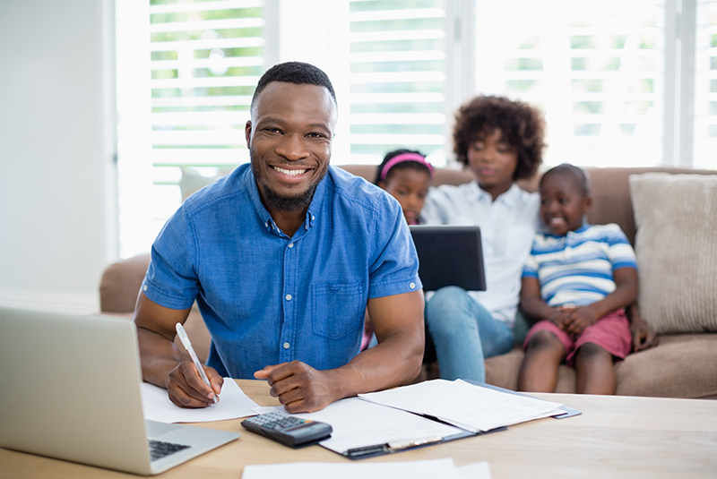 Man in the foreground working on a computer with a woman and two children in the background sitting on a couch