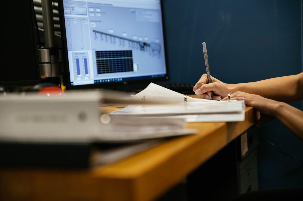 A person writes notes at a desk while reviewing information displayed on a computer screen.