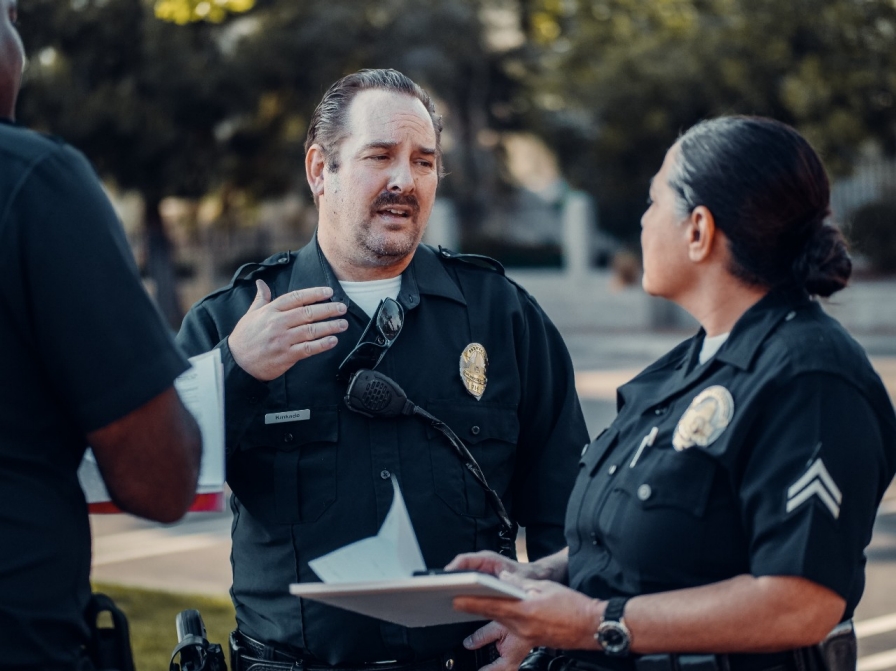 In an outdoor area, a police officer speaks to another officer who is holding a pad of paper.