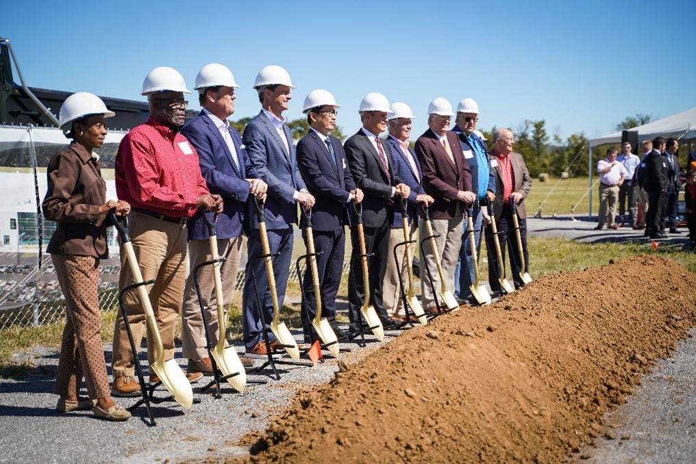 A group of people wearing hard hats stand in a line holding shovels at a groundbreaking event.