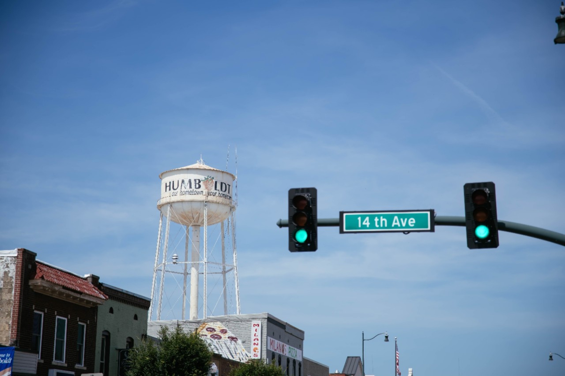 Water tower rises above downtown buildings near a traffic light and a street sign reading “14th Ave.”