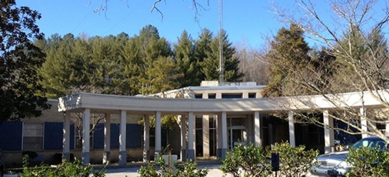 Single-story building with a covered entrance supported by columns, surrounded by trees and parked vehicles.