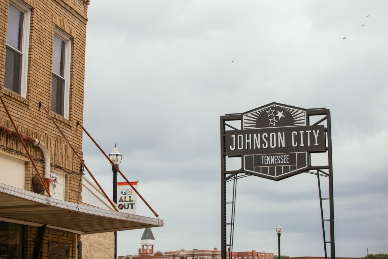 Downtown street scene with a large sign reading “Johnson City, Tennessee” beside brick buildings and streetlights under a cloudy sky.