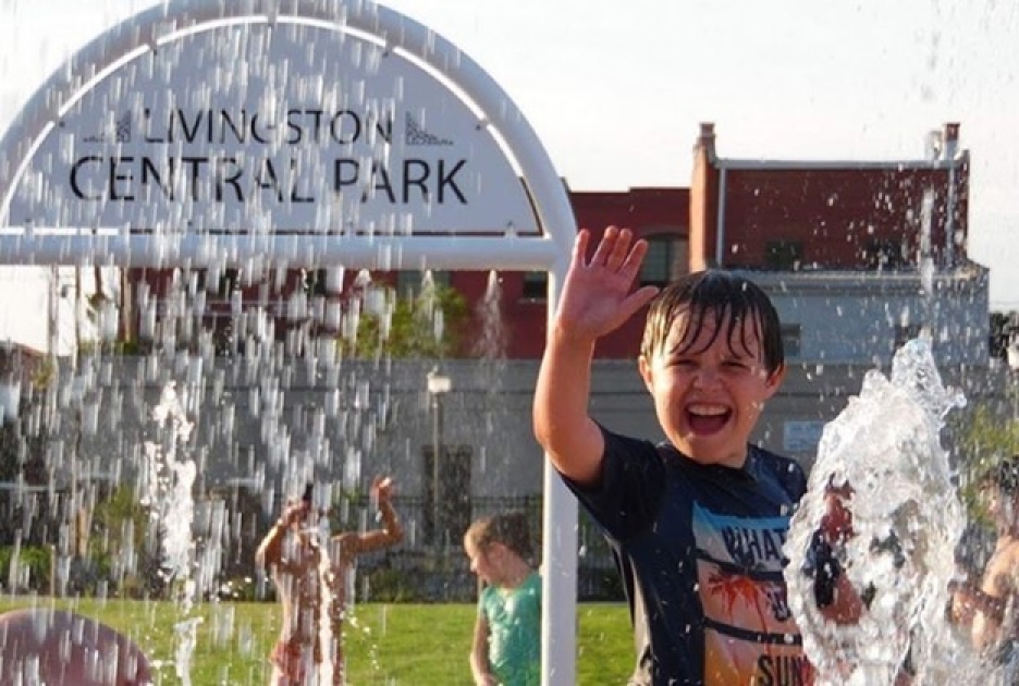 A child plays in a splash fountain at a public park while water sprays overhead.